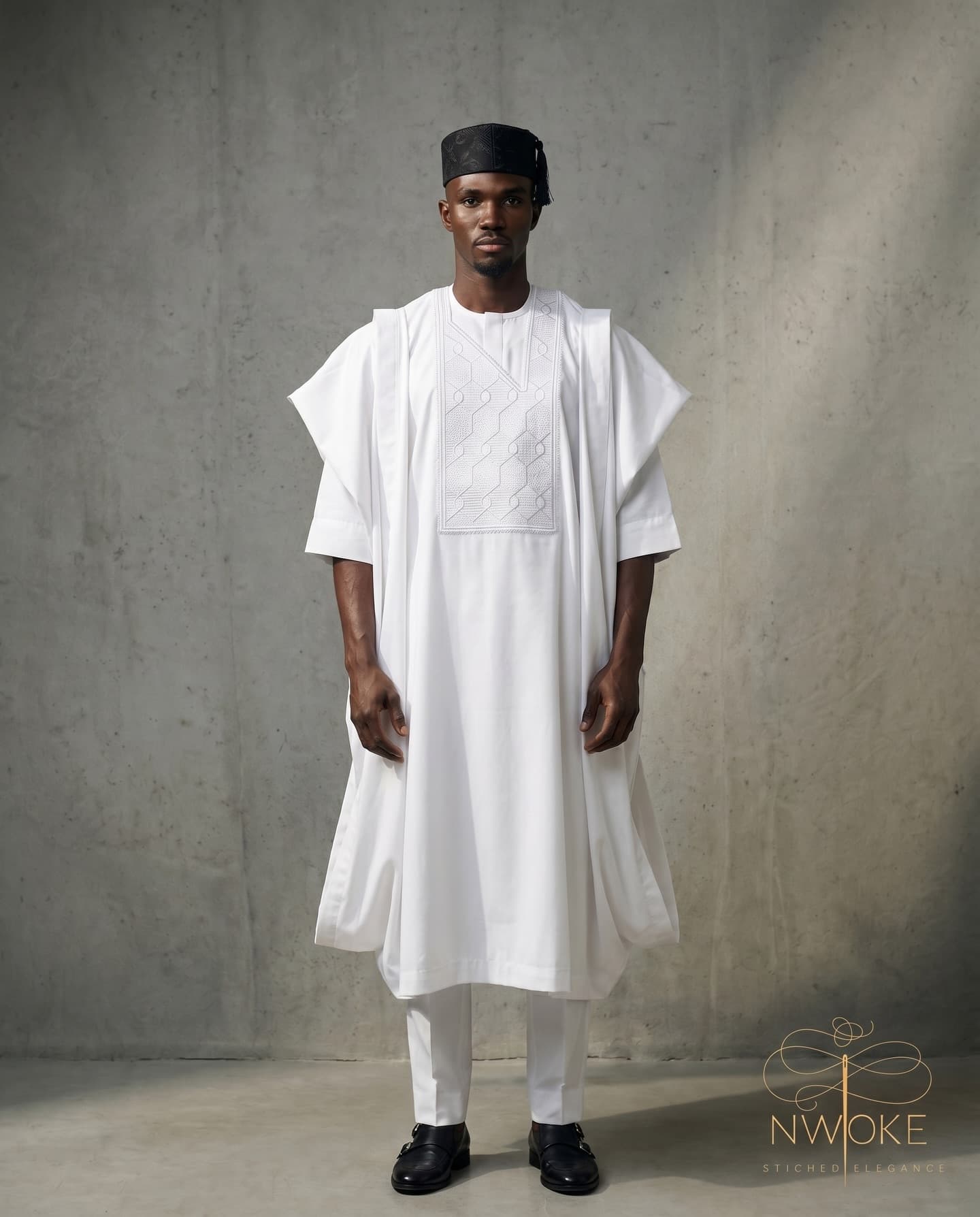 A model standing in the all-white NWOKÉ agbada and a dark fila against a grey backdrop