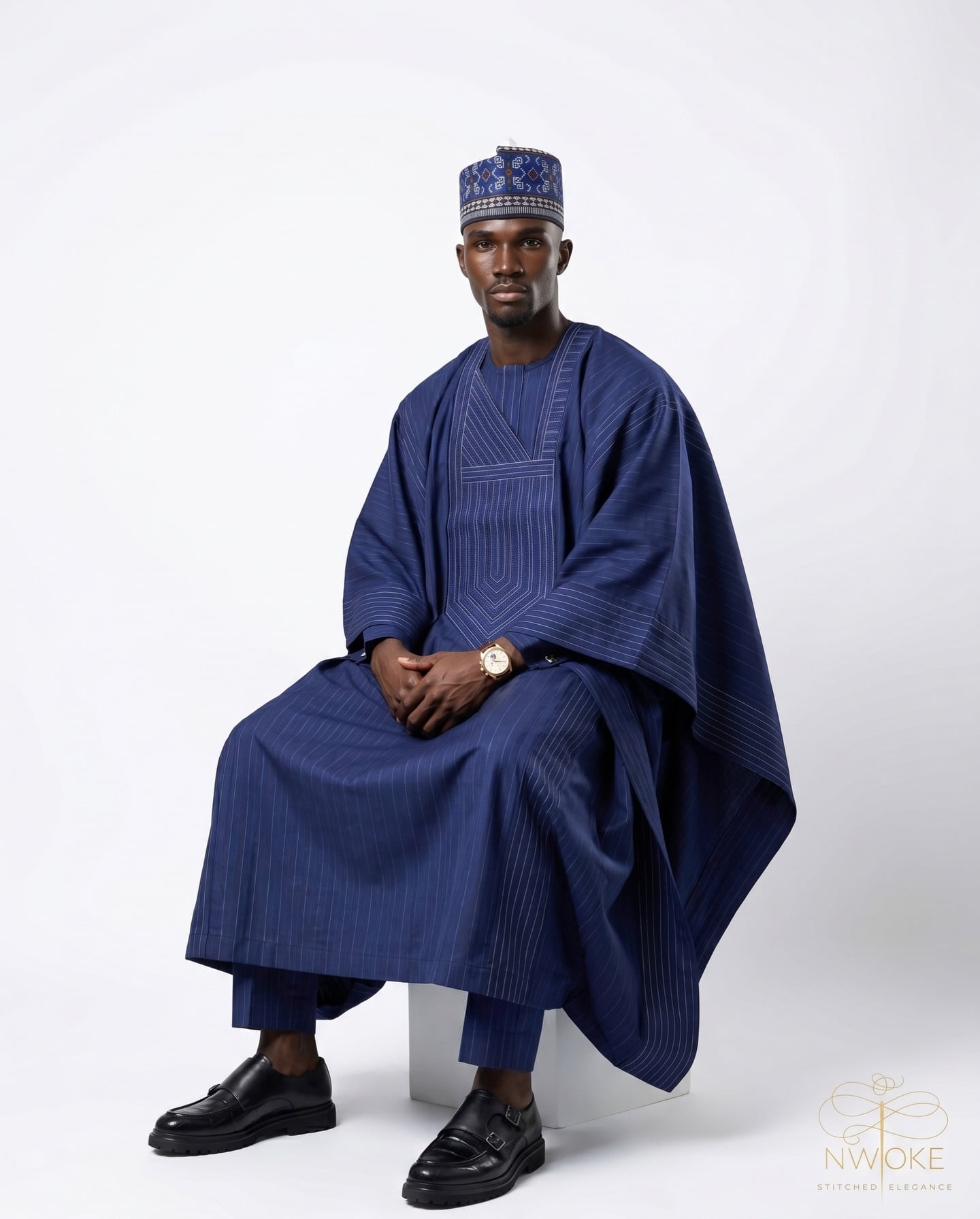 A man seated on a white block, wearing the NWOKÉ Midnight Agbada in deep indigo with a matching embroidered fila